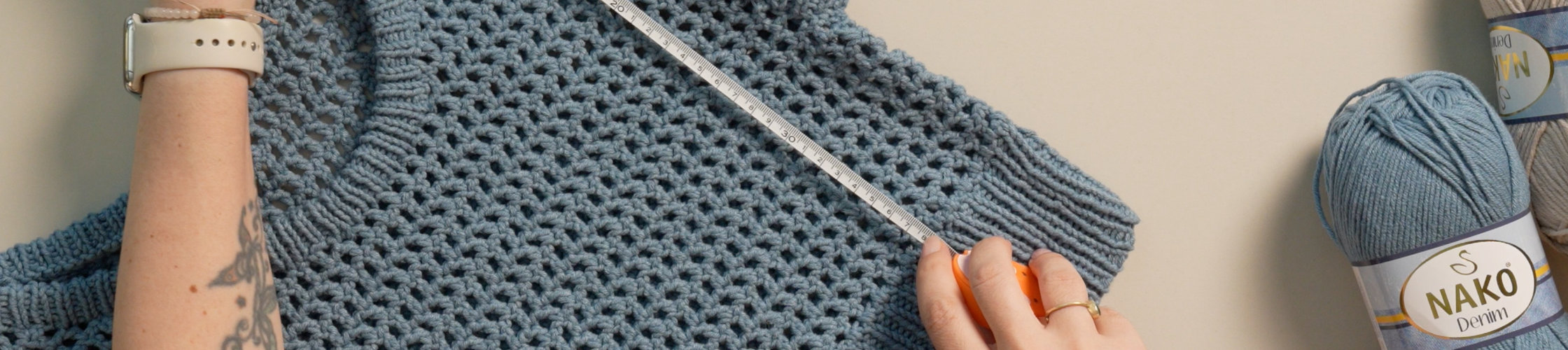 Hands measuring a knitted NAKO garment beside yarn skeins